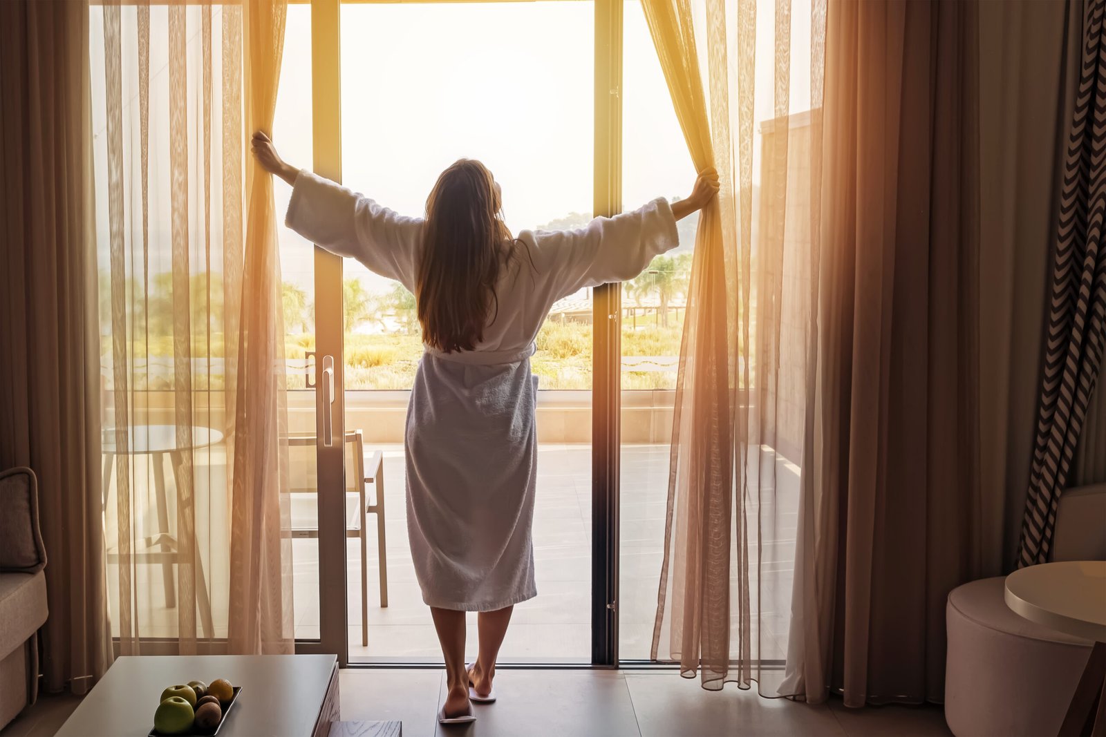 Young woman wearing white bathrobe opening curtains in luxury hotel room enjoying sun in the morning.